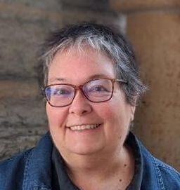 Picture: Photo of a white woman from the waist up, standing against a grayish stone building. She is fat, white, and smiling, with short, graying, dark hair and dark-rimmed glasses. She's wearing a black blouse under a denim jacket.